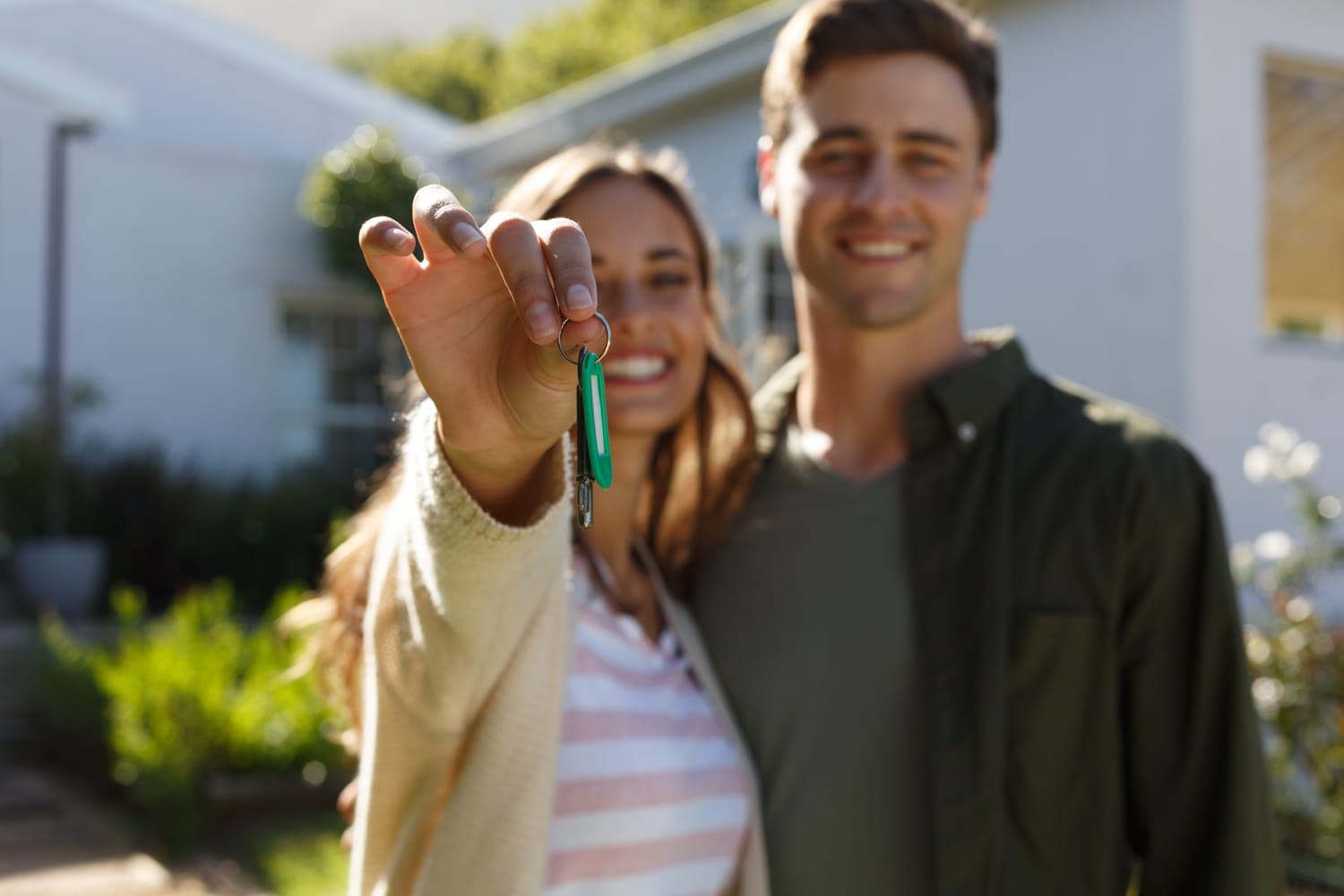 A happy couple standing in front of their new home, holding the keys on a bright, sunny day.
